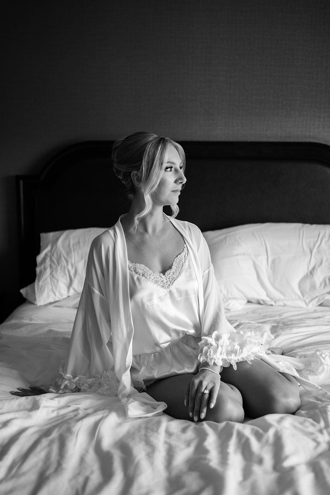 Black and white portrait of the bride sitting on a bed, looking thoughtfully out of the frame.